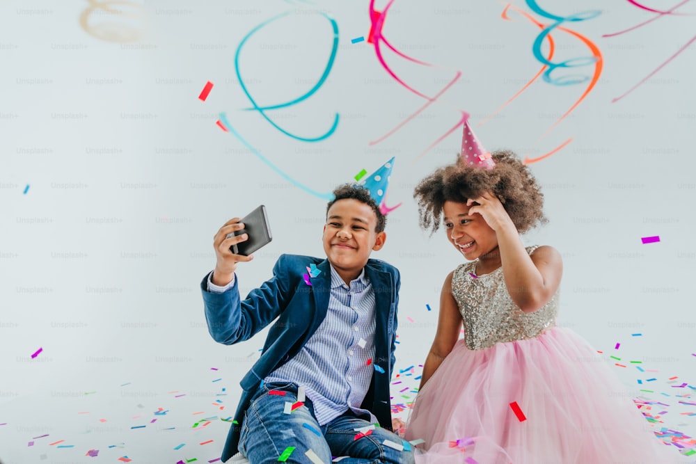 Happy girl and boy in party hats making selfie on smartphone on white background decorated with serpentine and confetti