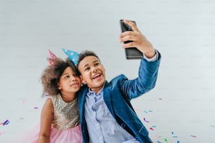 Menina e menino felizes em chapéus de festa fazendo selfie no smartphone no fundo branco decorado com serpentine e confete