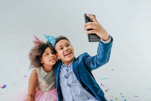 Happy girl and boy in party hats making selfie on smartphone on white background decorated with serpentine and confetti