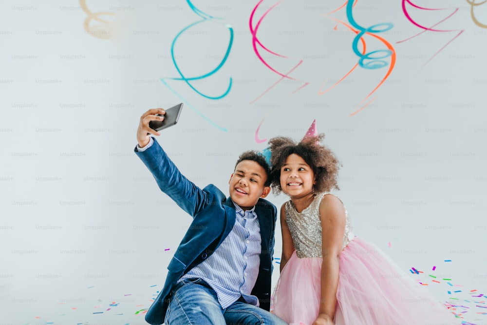 Glückliches Mädchen und Junge in Partyhüten machen Selfie auf Smartphone auf weißem Hintergrund, der mit Serpentin und Konfetti verziert ist