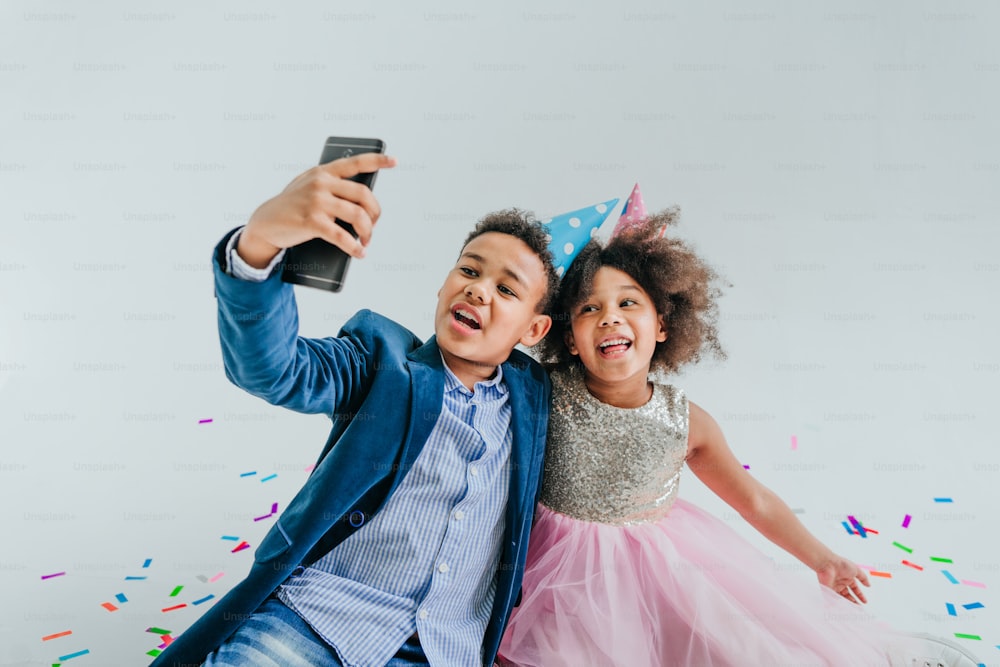Glückliches Mädchen und Junge in Partyhüten machen Selfie auf Smartphone auf weißem Hintergrund, der mit Serpentin und Konfetti verziert ist