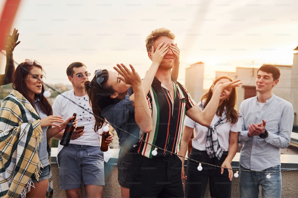 Playing game. Man's eyes covered by hands. Group of young people in casual clothes have a party at rooftop together at daytime.
