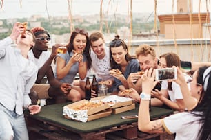 Making photo. Delicious pizza. Group of young people in casual clothes have a party at rooftop together at daytime.