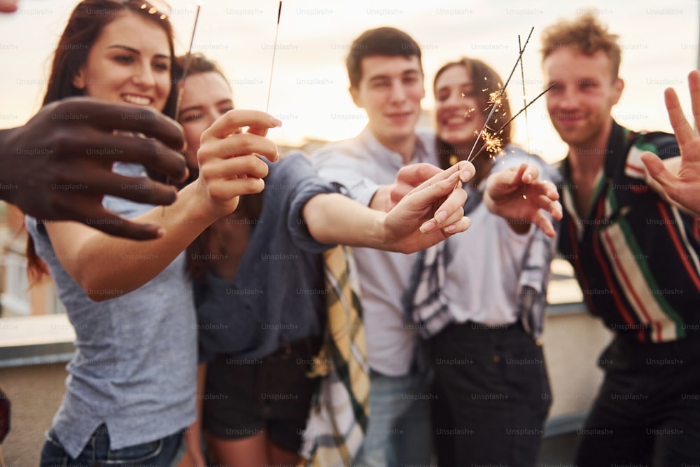 With sparklers in hands. Group of young people in casual clothes have a party at rooftop together at daytime.