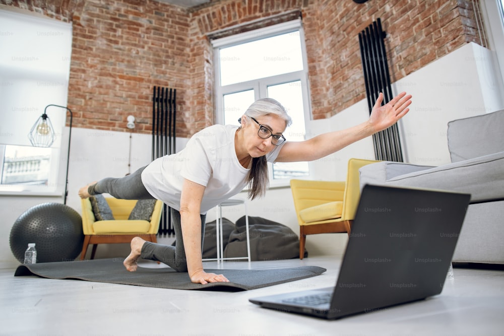 Retired lady watching online tutorial on laptop while doing yoga poses at home. Senior woman in activewear balancing on yoga mat on one knee and one hand.
