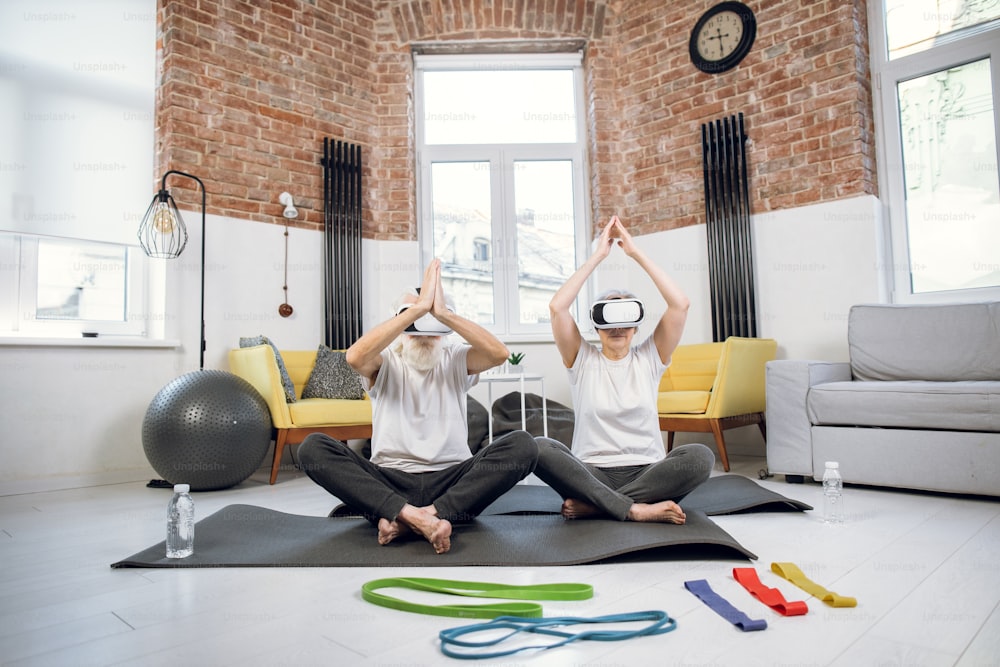 Retired man and woman in sport clothes meditating at home in VR headset. Senior couple practising yoga using modern technology.