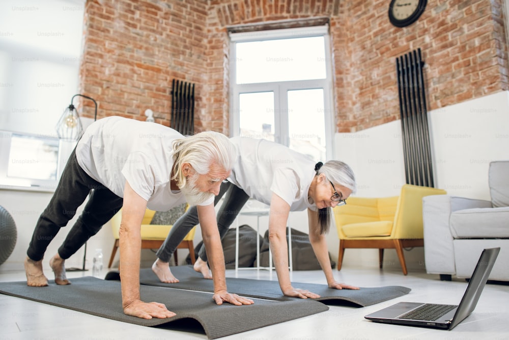 Mature man and woman in sport clothes doing downward dog pose while watching online training on wireless laptop. Concept of togetherness, active retirement and technology.