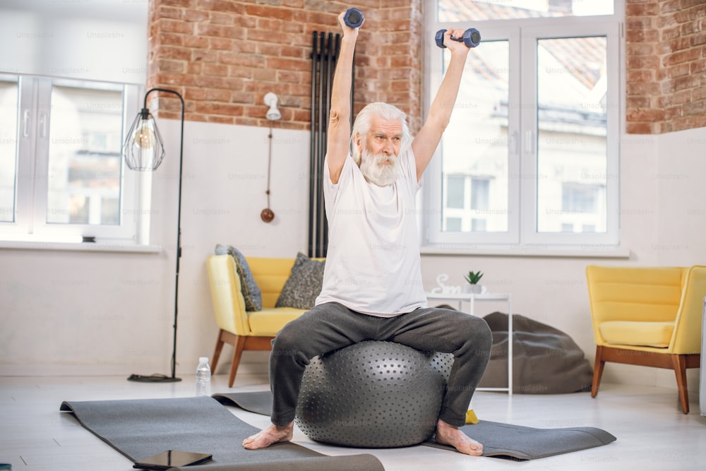 Mature man dressed in sport clothes using dumbbells and fitball for workout at home. Grey haired male sitting and holding hands up. Concept of healthy lifestyles.