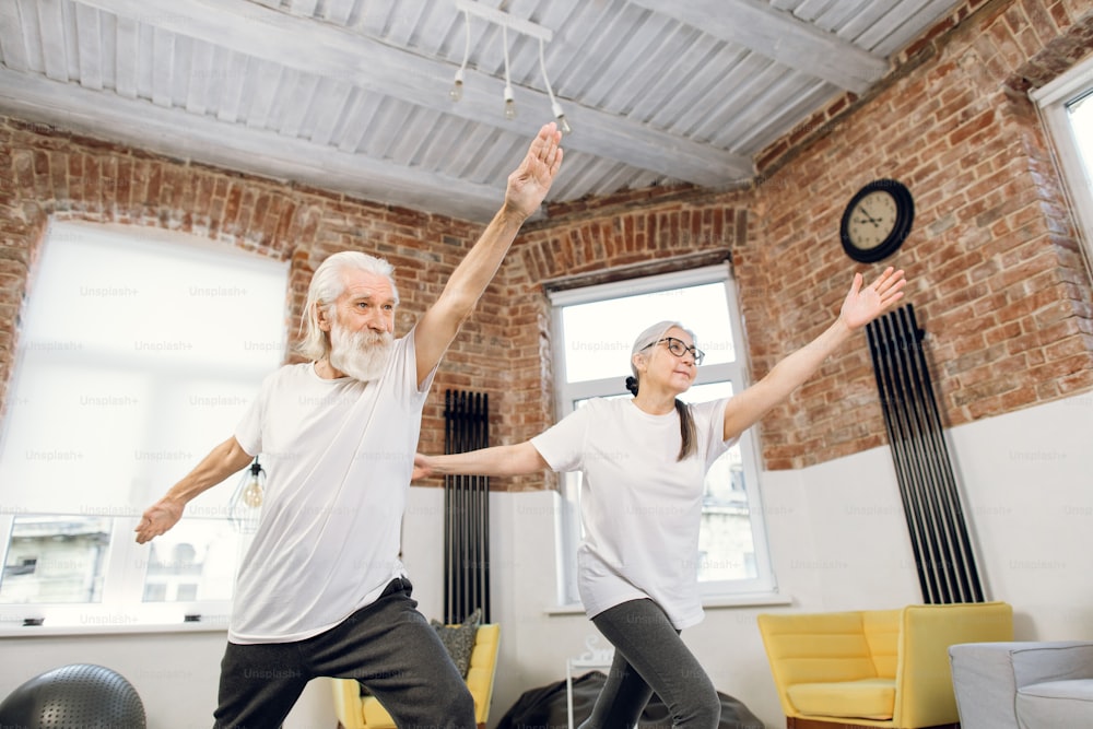 Zwei ältere Menschen in Sportkleidung stehen in Yoga-Asana-Pose und schauen sich das Online-Tutorial auf einem modernen Laptop an. Familientraining zu Hause. Aktiver Ruhestand.