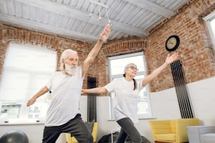 Two elderly people in sport clothes standing in yoga asana pose while watching online tutorial on modern laptop. Family training at home. Active retirement.