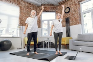 Full length portrait of elderly man and woman standing on yoga man and holding one hand up and one hand down. Modern laptop lying on floor. Active lifestyles.