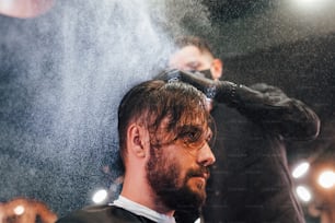 Young bearded man sitting and getting haircut in barber shop by guy in black protective mask.