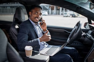 Smiling african man in business suit sitting in modern car with laptop on knees and talking on mobile phone. Young male showing thumb up and looking at camera.