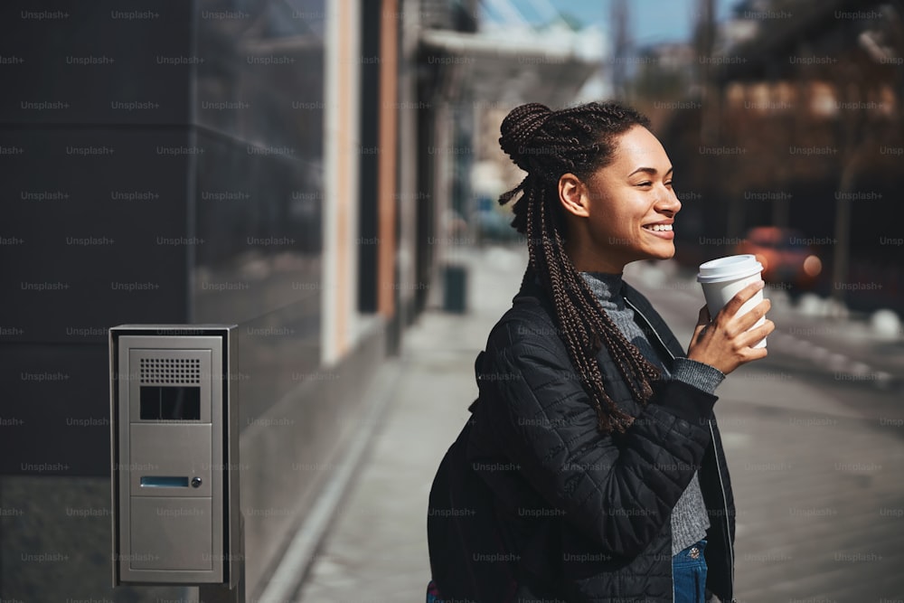 Young Afro American lady smiling in the street while drinking coffee near the building