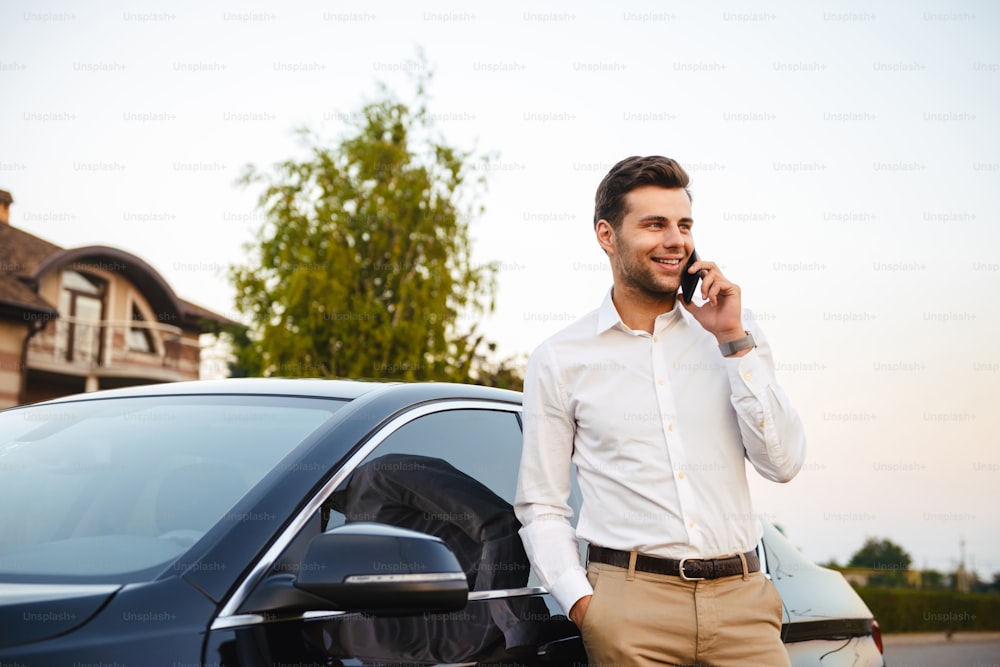 Portrait of handsome businessman wearing suit standing near his luxury black car and talking on mobile phone