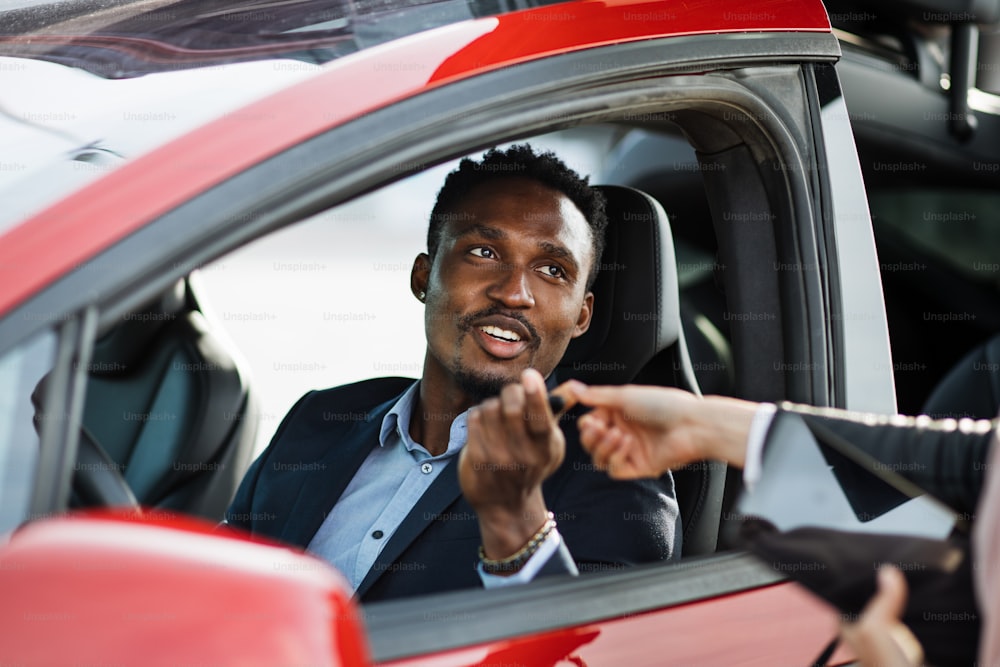 Uomo africano felice seduto all'interno della sua nuova auto elettrica mentre prende le chiavi dal rivenditore femminile. Uomo d'affari di successo in giacca e cravatta che acquista un veicolo ecologico.
