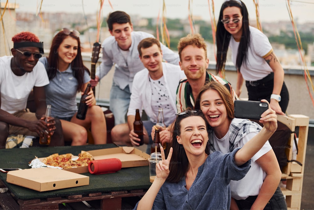 Girl making selfie. With delicious pizza. Group of young people in casual clothes have a party at rooftop together at daytime.
