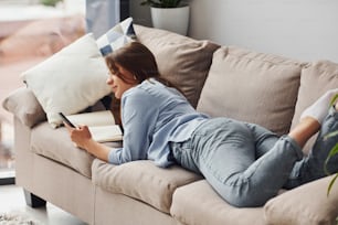 Bela mulher jovem em jeans e camisa azul deitada no sofá com telefone na mão.