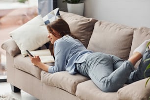 Beautiful young woman in jeans and blue shirt lying down at sofa with phone in hand.