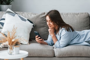 Beautiful young woman in jeans and blue shirt lying down at sofa with phone in hand.