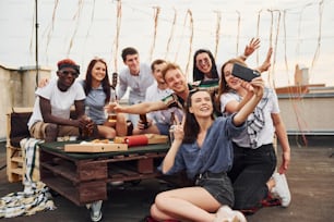 Girl making selfie. With delicious pizza. Group of young people in casual clothes have a party at rooftop together at daytime.