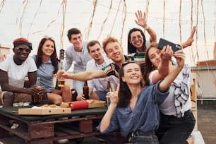 Girl making selfie. With delicious pizza. Group of young people in casual clothes have a party at rooftop together at daytime.