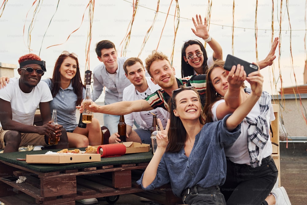 Mädchen macht Selfie. Mit leckerer Pizza. Eine Gruppe junger Leute in Freizeitkleidung feiert tagsüber gemeinsam auf dem Dach.