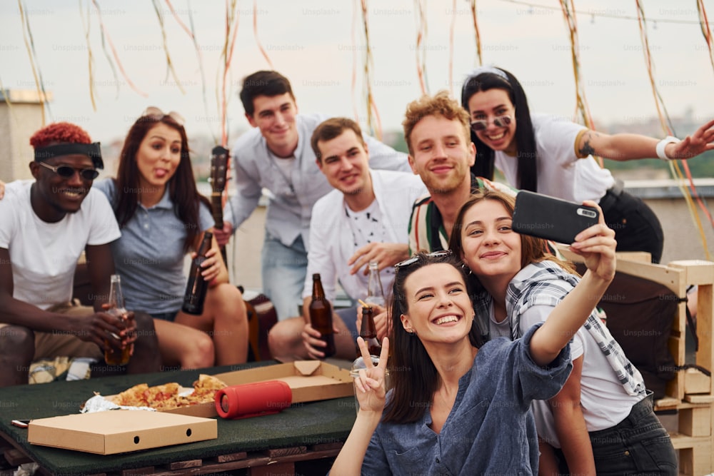 Ragazza che fa selfie. Con una pizza deliziosa. Un gruppo di giovani in abiti casual fa una festa insieme sul tetto durante il giorno.