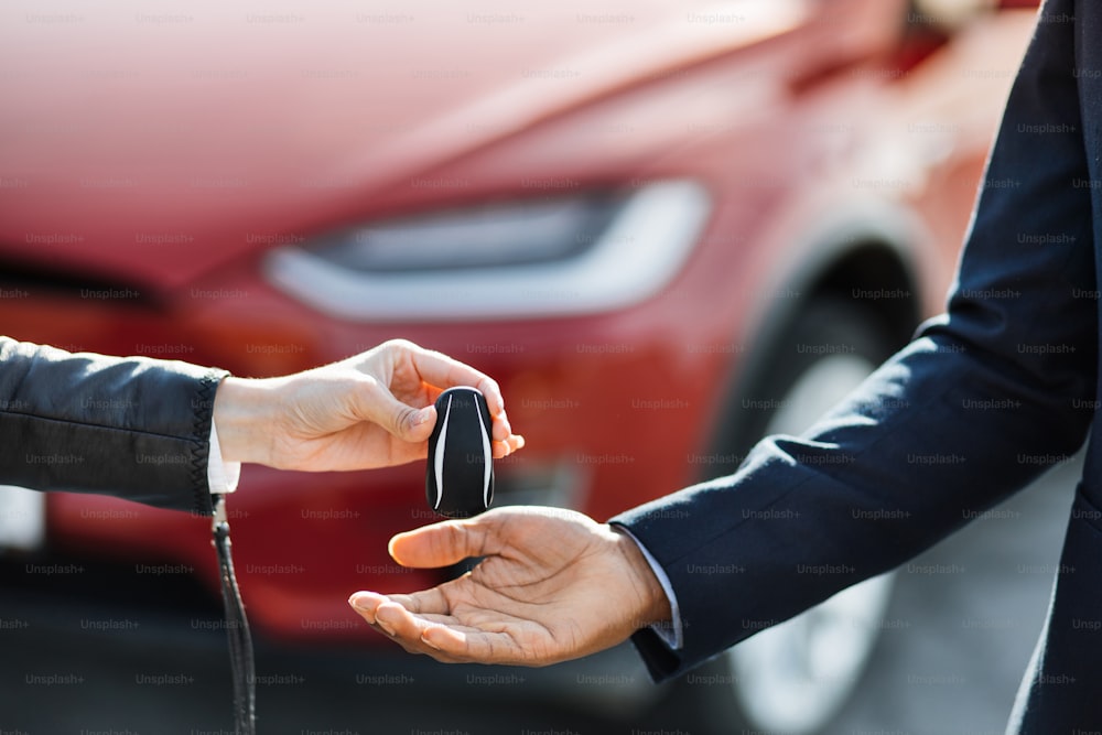 Primo piano del venditore femminile che presenta le chiavi dell'auto elettrica rossa al cliente maschile. Sfocatura dello sfondo dell'auto di lusso. Concetto di affare riuscito.
