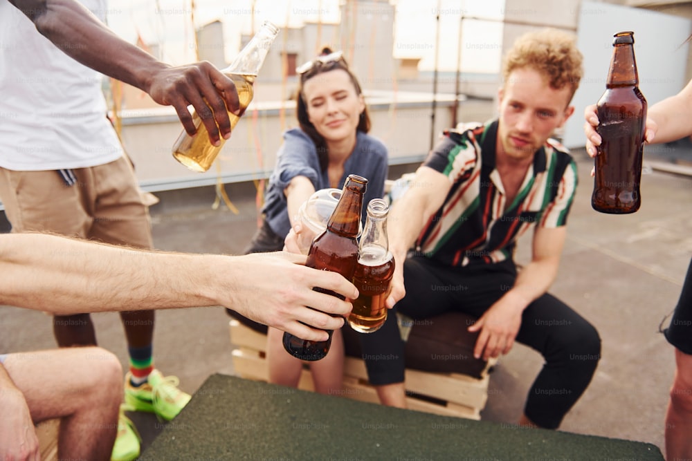 Doing cheers by bottles with beer. Group of young people in casual clothes have a party at rooftop together at daytime.