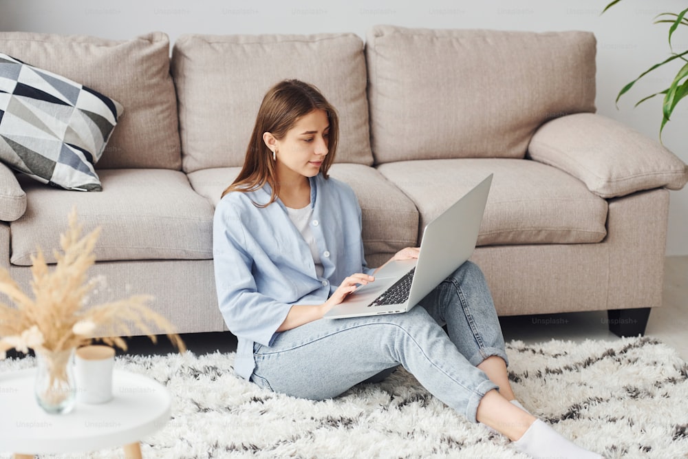 Beautiful young woman in jeans and blue shirt sits on the floor with laptop indoors at home.