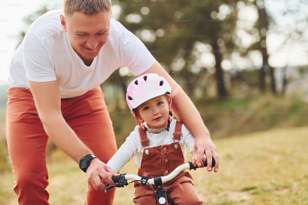 Father in white shirt teaching daughter how to ride bicycle outdoors.