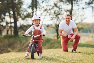 Père en chemise blanche apprenant à sa fille à faire du vélo à l’extérieur.