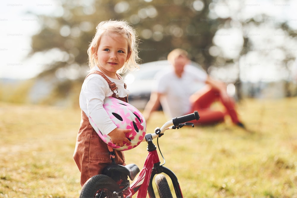 Father in white shirt sitting on the ground and his daughter standing in front of camera with her bike.