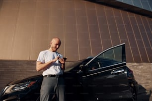 Glatzköpfiger Mann, der tagsüber mit dem Telefon in der Hand in der Nähe seines modernen schwarzen Autos im Freien steht.