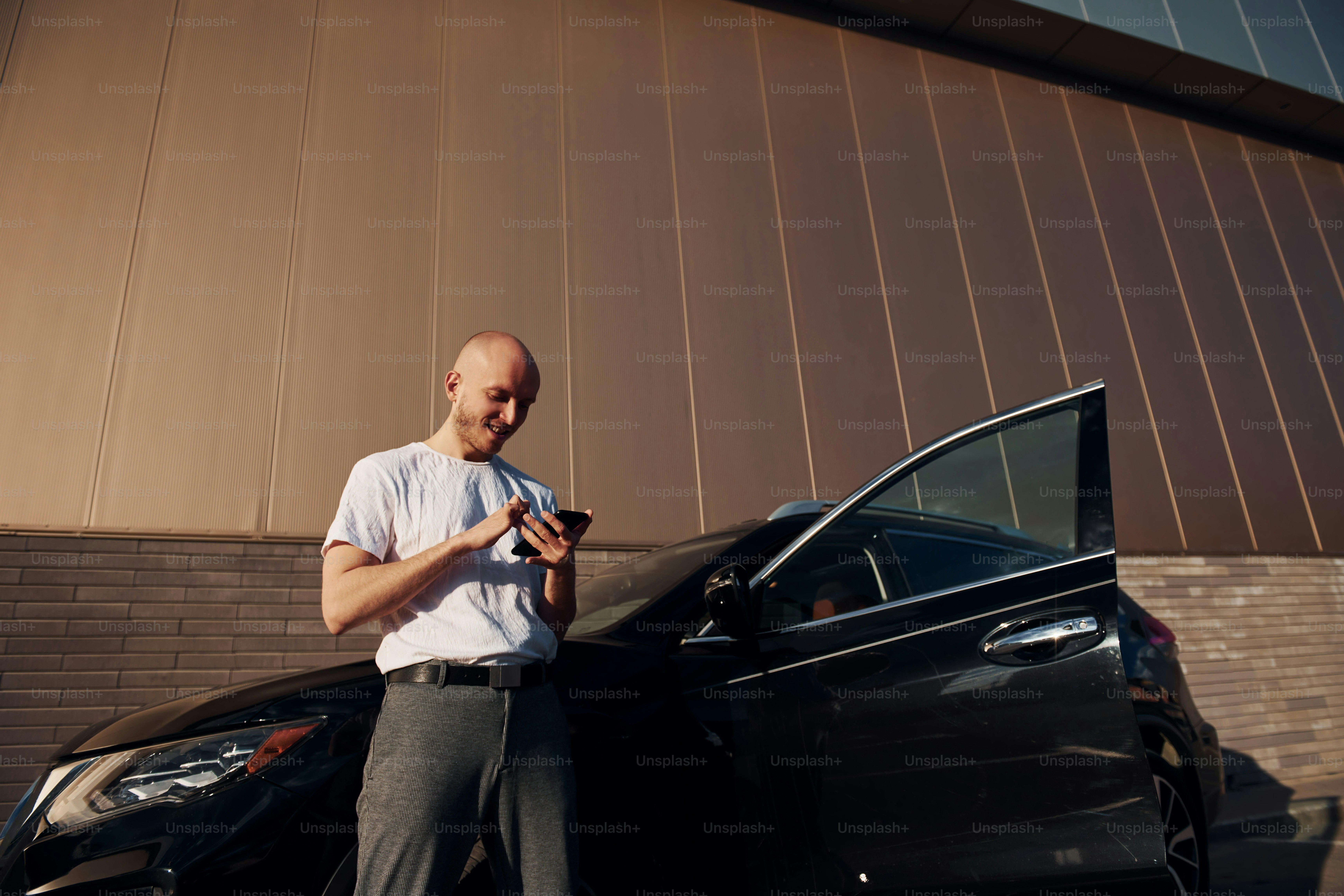 Bald man standing near his modern black car outdoors at daytime with phone in hands.