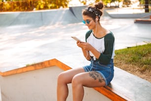 Photo of young smiling pretty woman sitting outdoors listening music with earphones using mobile phone.