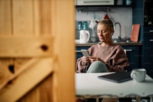 Pretty senior woman sitting in the kitchen at home and using phone for social media, listening to the music or video call