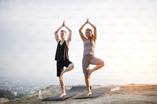 Full length portrait of multi ethnic couple in sport clothes standing on one leg and holding hands above head, Vrksasana, Tree pose, during yoga practice outdoors. Concept of healthy lifestyles.