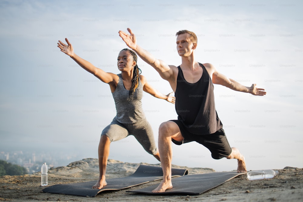Caucasian man and african american woman dressed in activewear standing in  with bare feet on yoga mat and exercising together with arms outstretched.  View of big city among clouds on background. photo –
