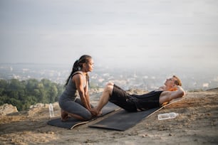 Starker kaukasischer Mann, der Bauchknirschen auf Yogamatte macht, während eine afrikanische Frau seine nackten Füße hält. Multirassisches aktives Paar, das Freizeit für Aktivitäten im Freien verbringt.