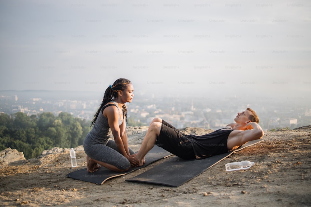 Hombre caucásico fuerte haciendo abdominales abdominales en una colchoneta de yoga mientras una mujer africana sostiene sus pies descalzos. Pareja activa multirracial que pasa tiempo libre para actividades al aire libre.