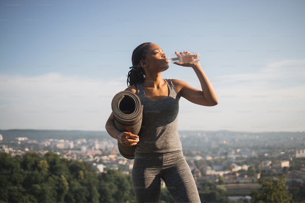 Attractive firtness woman with black skin drinking fresh water from bottle after training outdoors. Young female in sport clothes refreshing after workout.