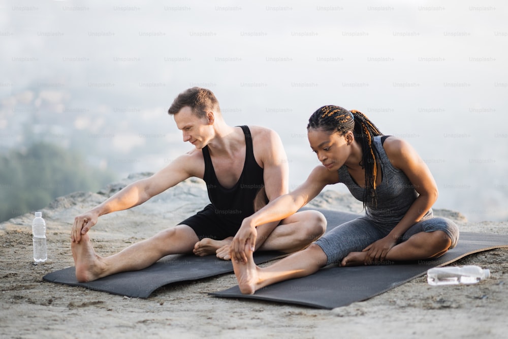 African american woman and caucasian man stretching legs on yoga mat on top of high rock. Young couple in activewear exercising together on fresh air.