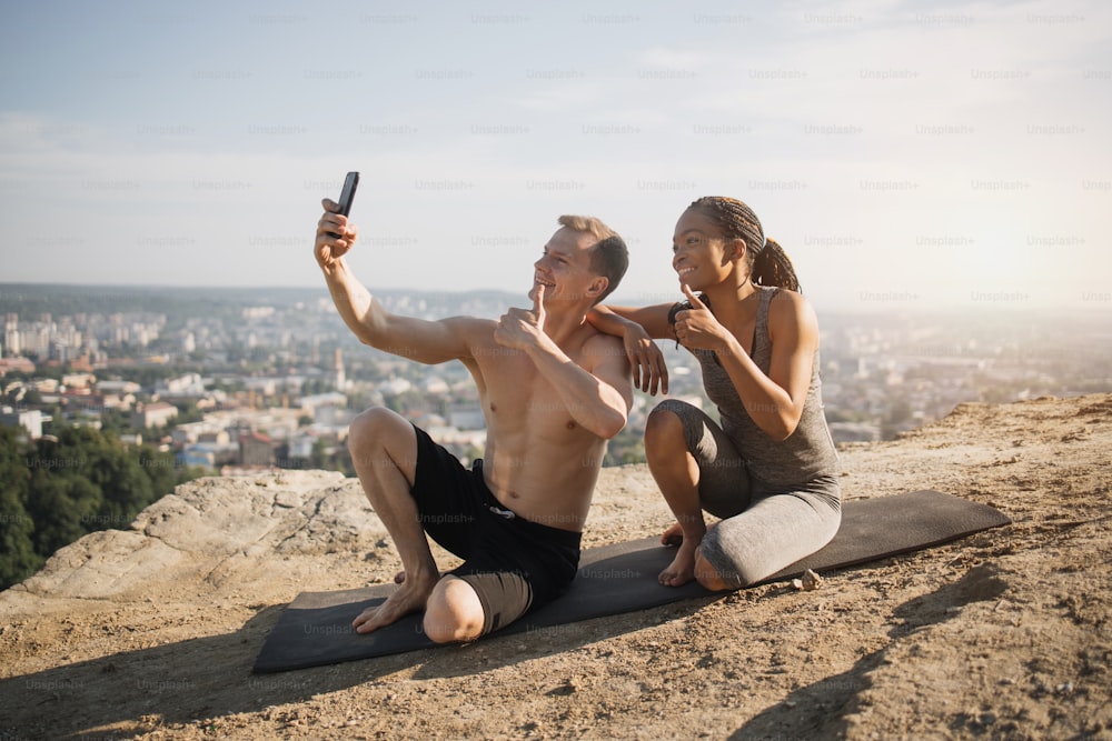Happy multiracial couple in sport outfit sitting together on yoga mat and using smartphone for taking selfie. Man and woman relaxing after workout with gadget in hands, showing thumbs up.