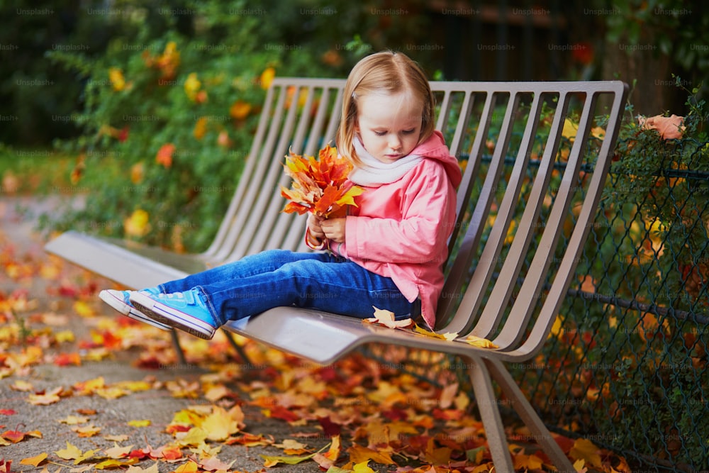 Adorable niña pequeña sentada en el banco y recogiendo hojas caídas en el parque de otoño. Niño feliz disfrutando del día de otoño en París, Francia. Actividades al aire libre para niños