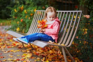 Adorable niña pequeña sentada en el banco y recogiendo hojas caídas en el parque de otoño. Niño feliz disfrutando del día de otoño en París, Francia. Actividades al aire libre para niños