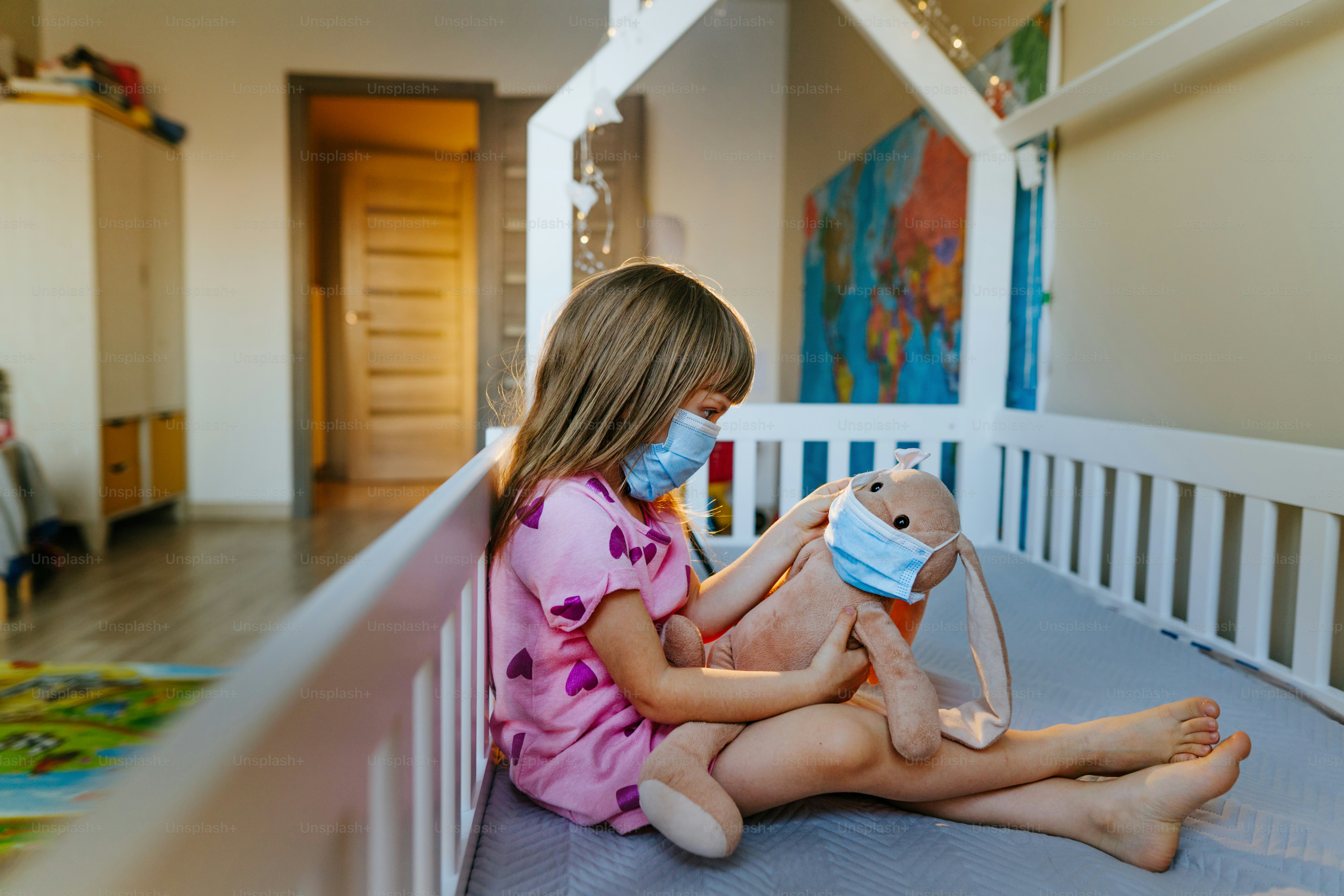 Kleines 4-jähriges Mädchen mit Schutzmaske spielt mit ihrem Plüschtierhasen, das auf dem Bett im Kinderzimmer sitzt. Neue Normalität nach pandemischem COVID-19-Konzept.