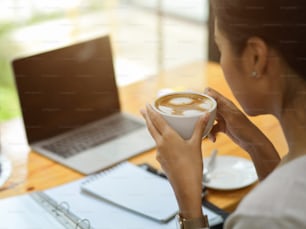 Close up female drink hot coffee at co-working space, businesswoman drink coffee while watch business strategy video, opened laptop, reports and coffee cup