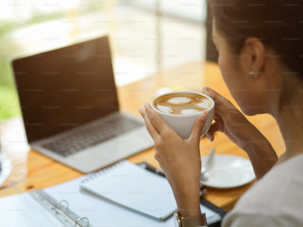 Nahaufnahme weiblich trinkt heißen Kaffee im Coworking Space, Geschäftsfrau trinkt Kaffee, während sie Business-Strategie-Video anschaut, geöffneter Laptop, Berichte und Kaffeetasse